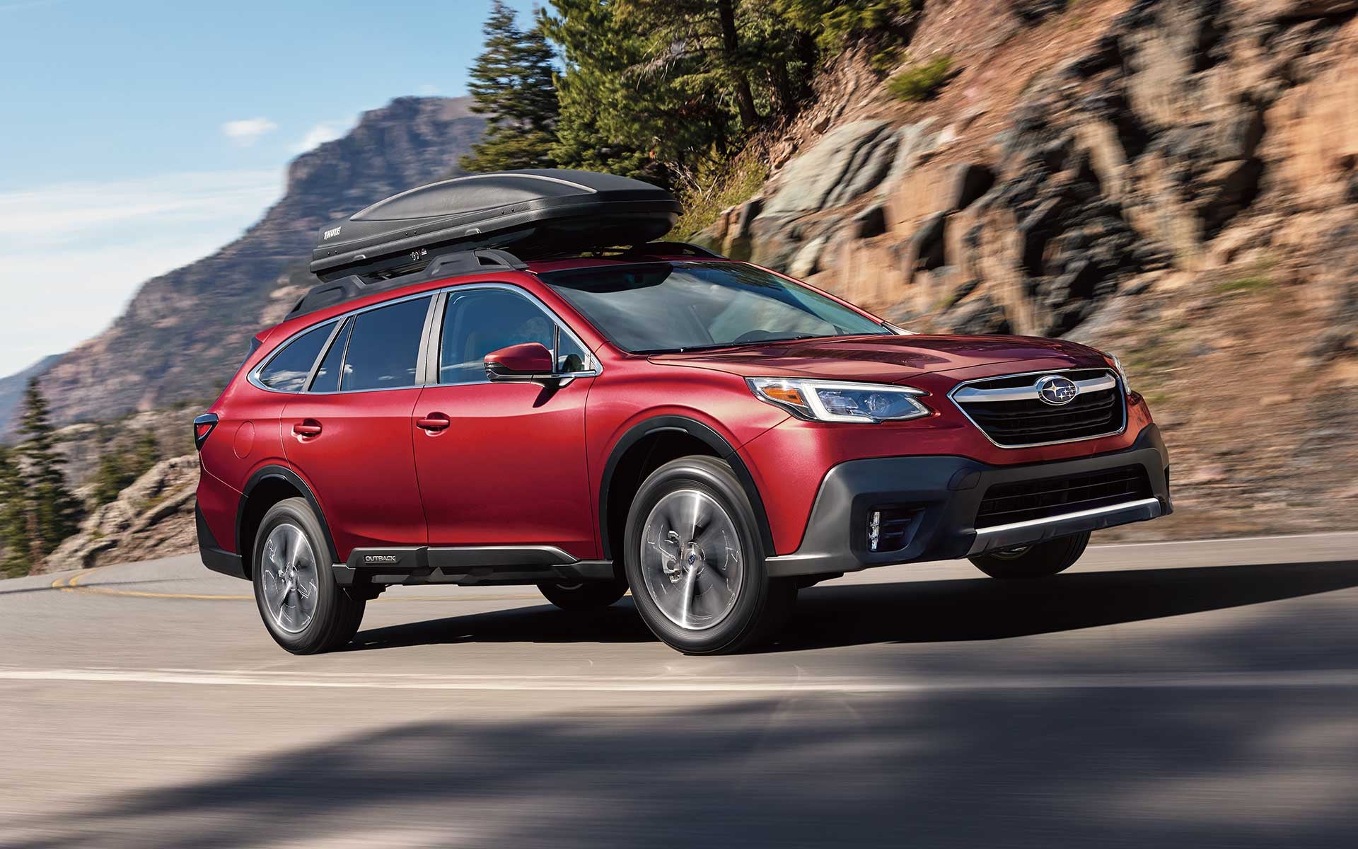 Red Subaru Outback with a black roof cargo box driving on a winding mountain road with rocky cliffs and trees.