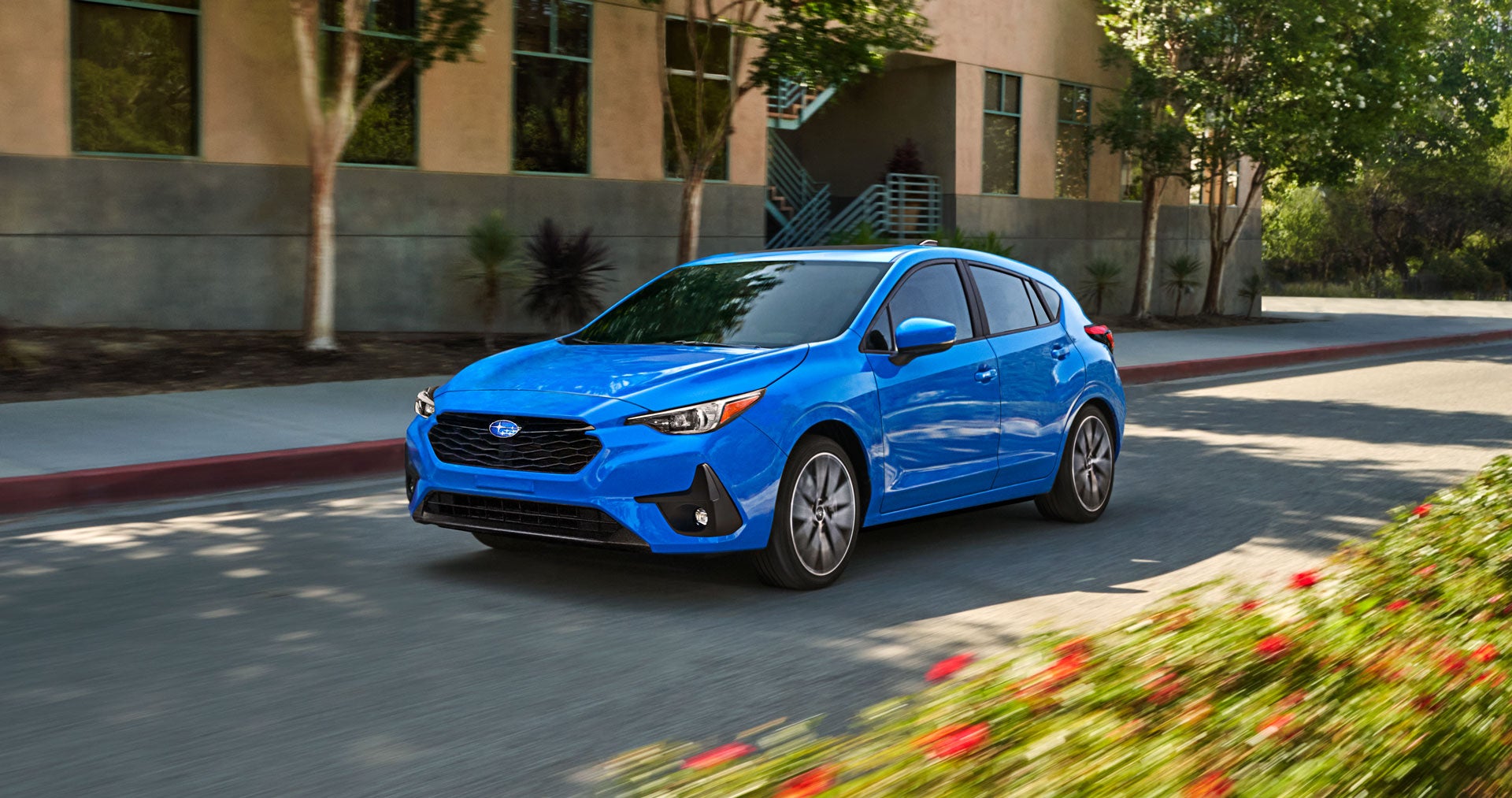 Bright blue hatchback car driving on a sunny street with blurred background buildings and trees.