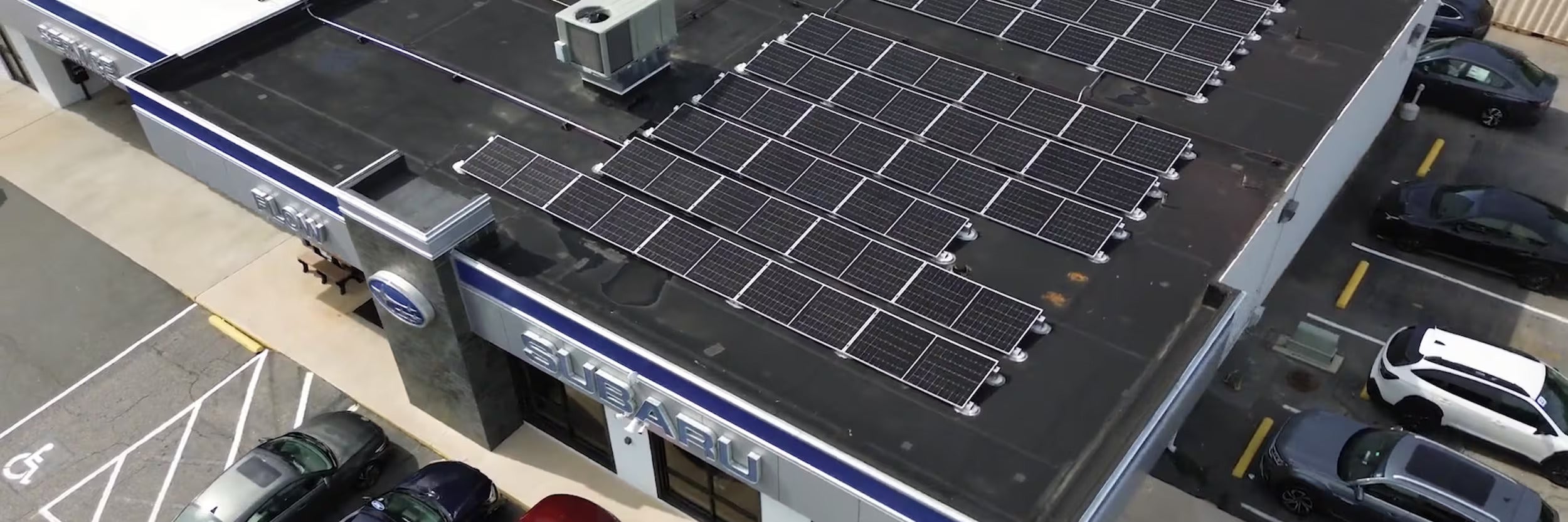 Aerial view of a Subaru dealership with many solar panels covering its flat roof and cars parked in the lot.