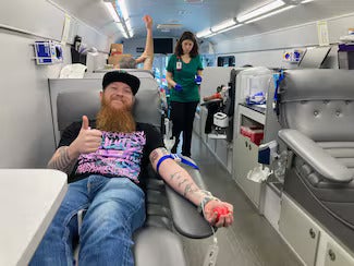 Man with beard and tattoos gives a thumbs up while donating blood in a mobile blood donation center.