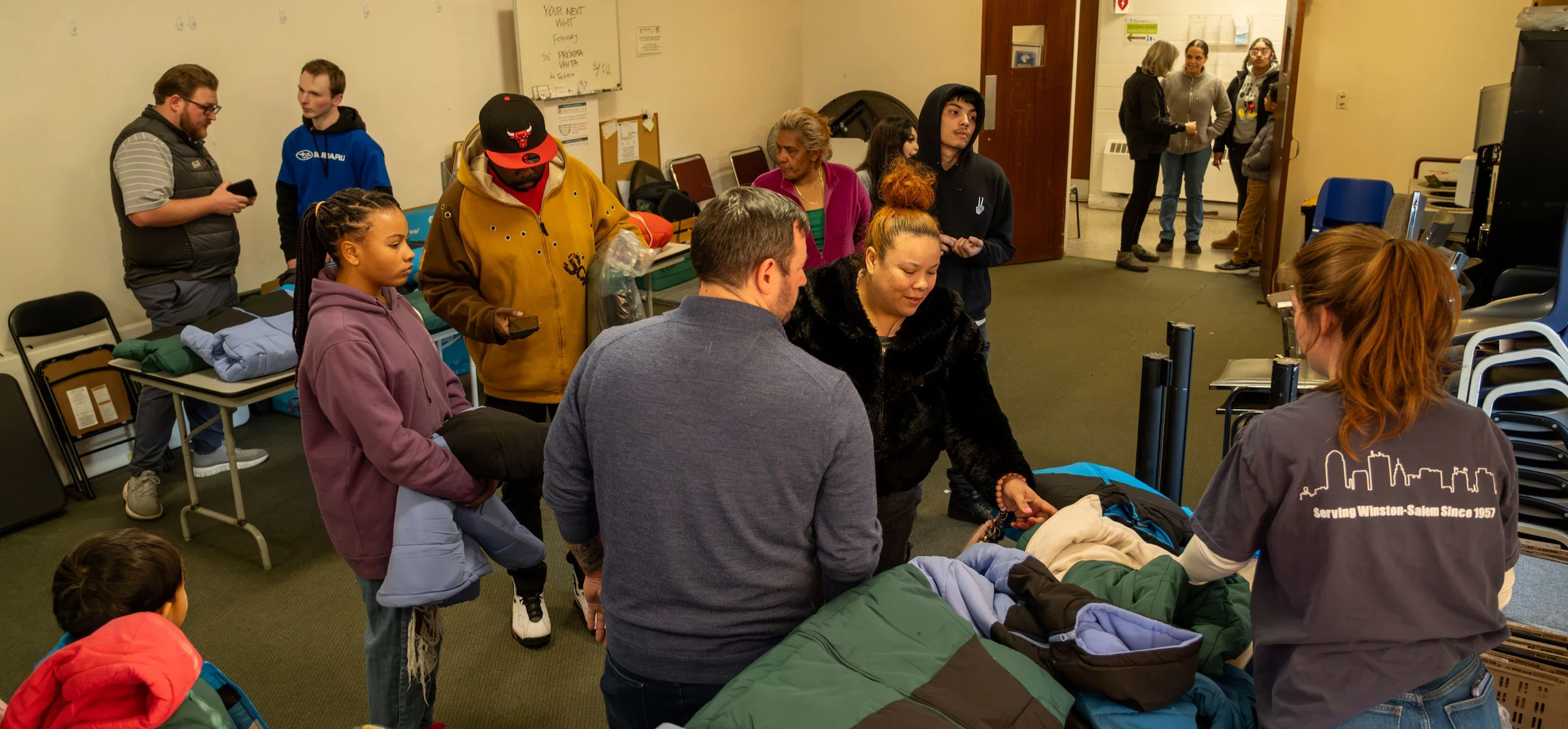 Diverse people gather in a room, some handling winter coats from tables, suggesting a community distribution event.
