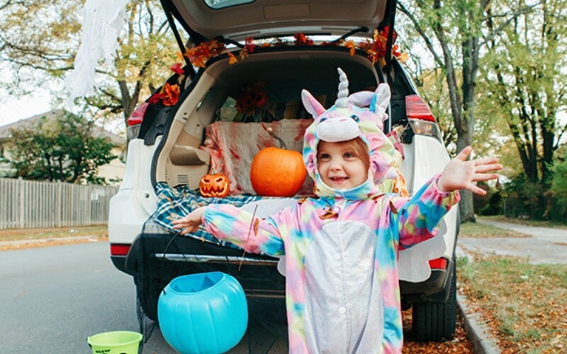 Happy child in a unicorn costume stands before a Halloween-decorated car trunk with pumpkins.