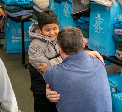 A happy child in a winter coat embraces an adult, surrounded by stacks of blankets and blue boxes.