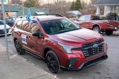 Red Subaru SUV with a blue flag on its roof rack and a sign on the passenger door, parked on a street.