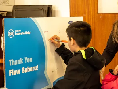 Boy writing on a Thank You Flow Subaru! sign, next to Subaru Loves to Help logo.