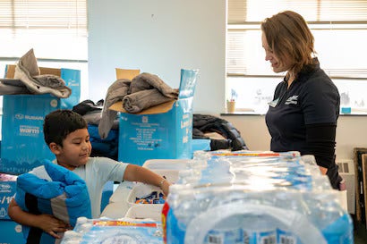 Young boy holding a blanket, reaching into a bin, as a smiling woman watches him at a Subaru Operation Warm event.